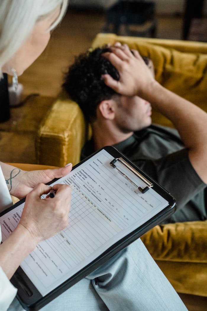 A therapist writes on a clipboard while a man holds his head, depicting a therapy session.